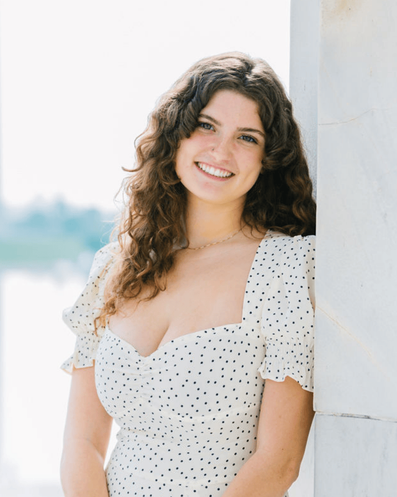 Portrait of a smiling woman with long curly hair wearing a white polka-dot dress, standing outdoors and leaning against a column.